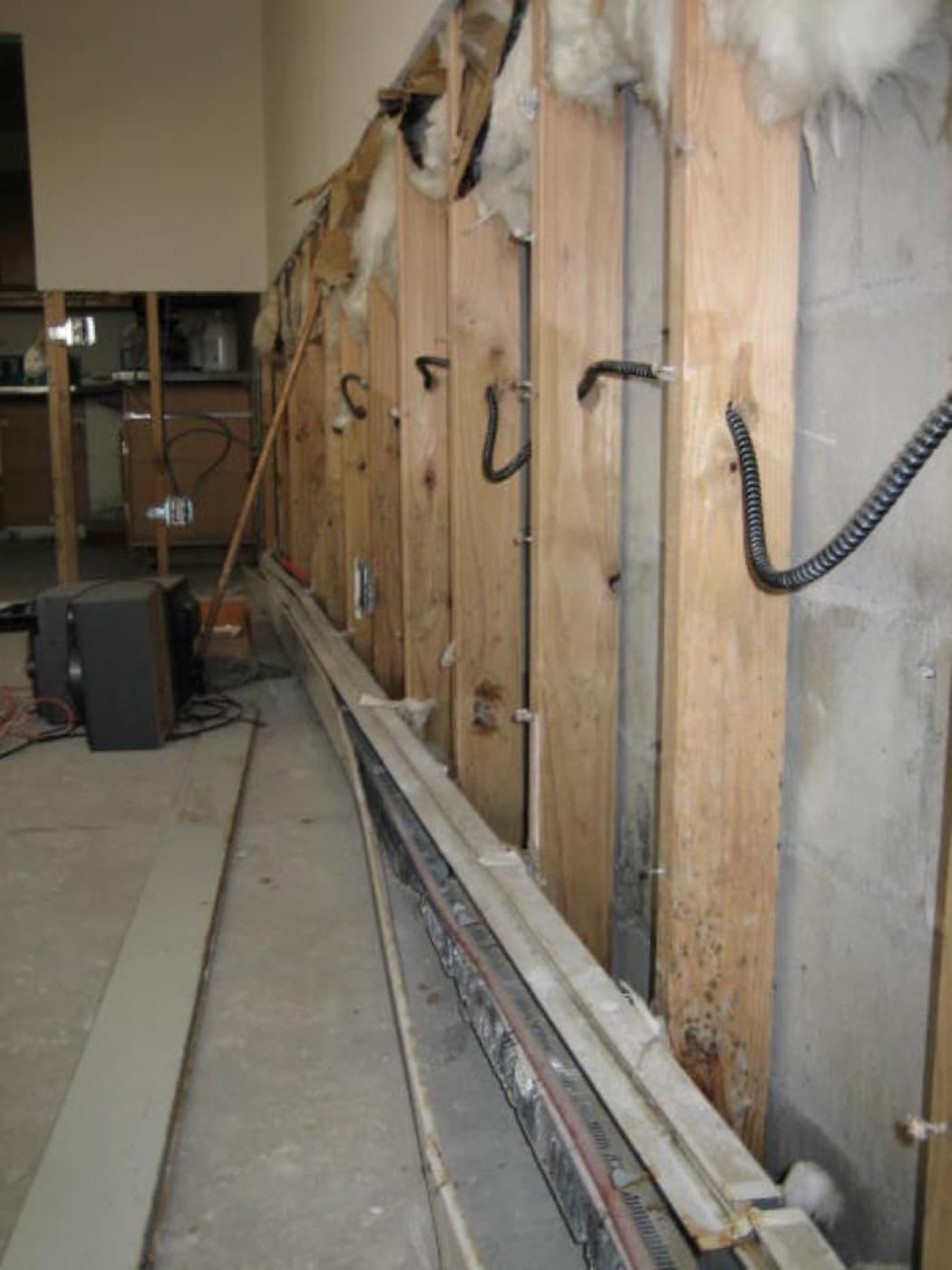 Interior wall with drywall and flooring removed, exposing wooden studs with mold growth and a drying fan running